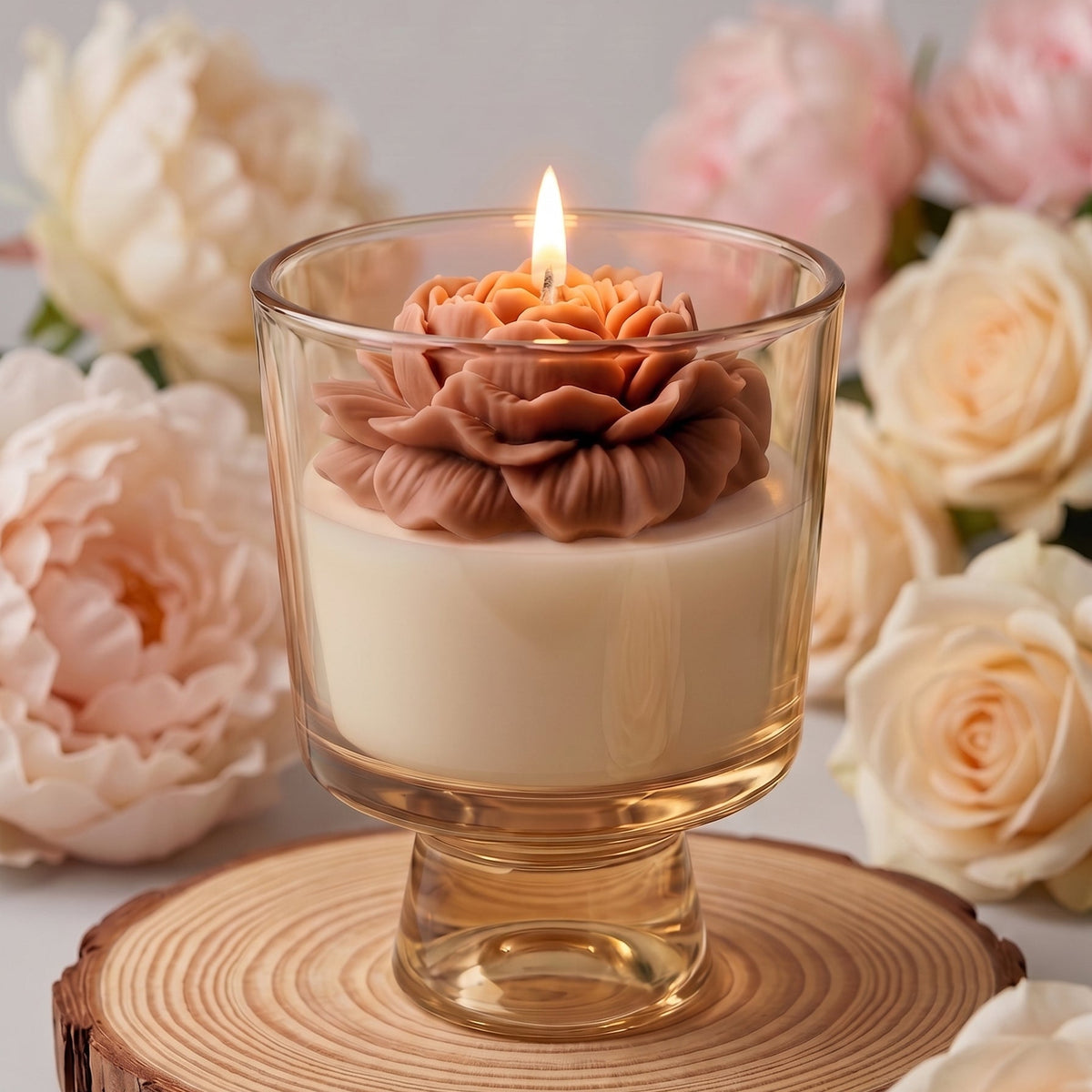 A lit candle in a clear glass pedestal cup sits on a round wood slice coaster, surrounded by soft pink and cream roses against a neutral gray background.