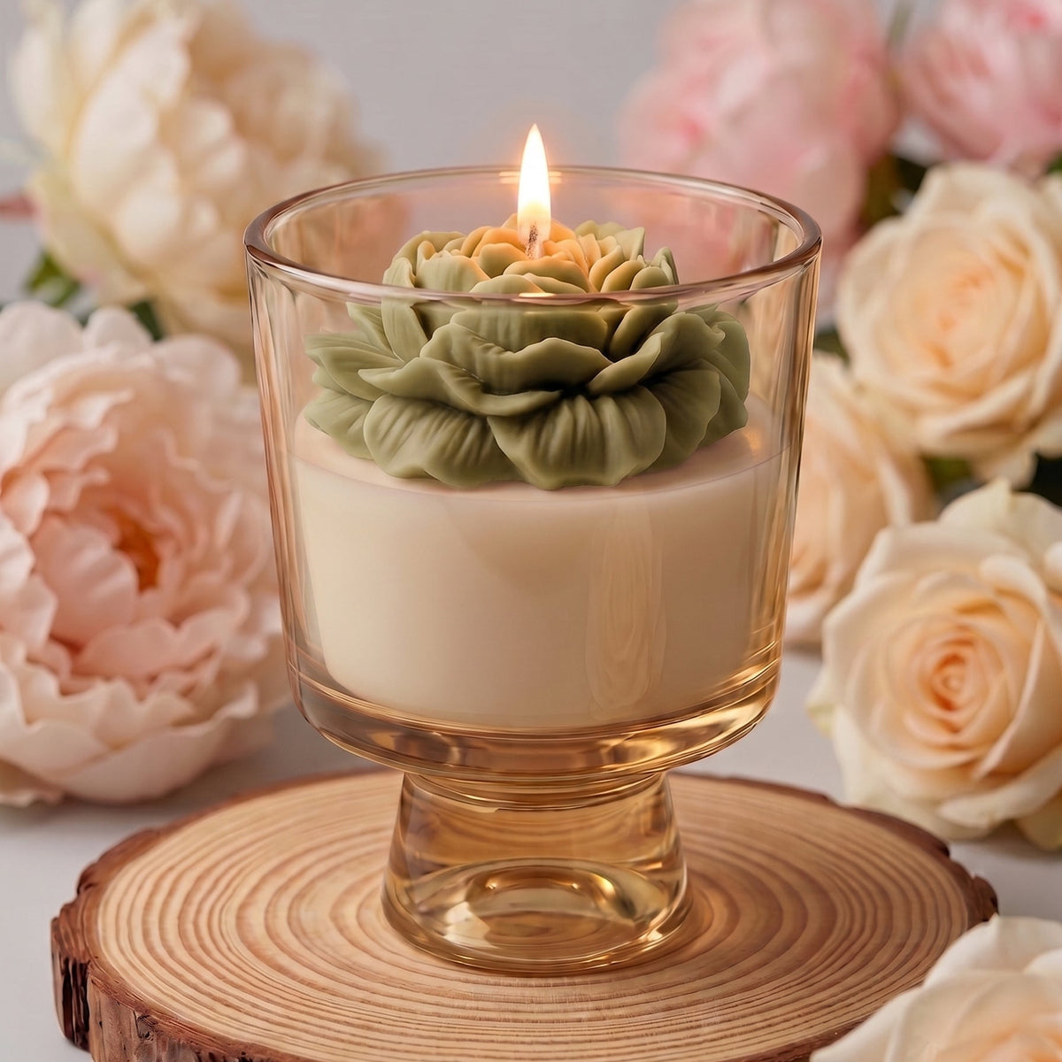 A lit candle in a clear glass pedestal holder sits on a round wood slice coaster, surrounded by soft pink and cream roses against a light gray background.