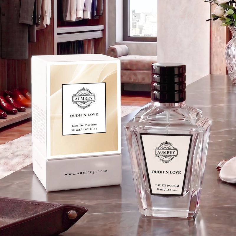 A clear glass perfume bottle labeled AUMBEY OUDH N LOVE stands beside its cream-colored box on a metallic tabletop in a styled interior setting.