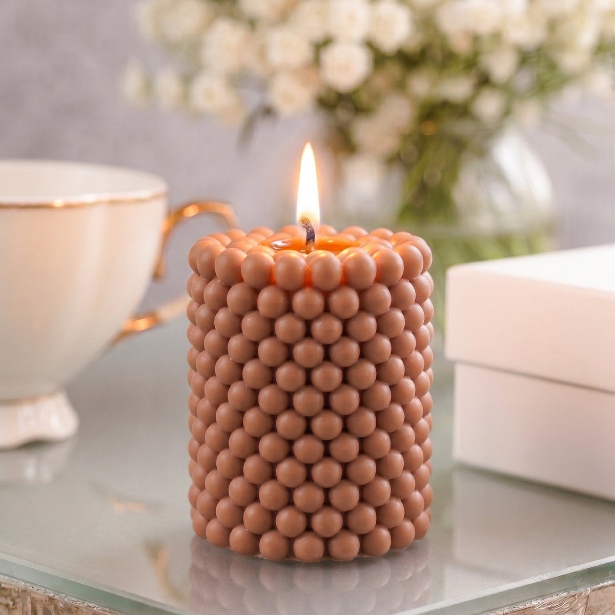 A lit cylindrical candle with a textured, beaded surface in a warm terracotta-brown color, styled on a glass table with a white teacup, white boxes, and a blurred bouquet of white flowers in the background.