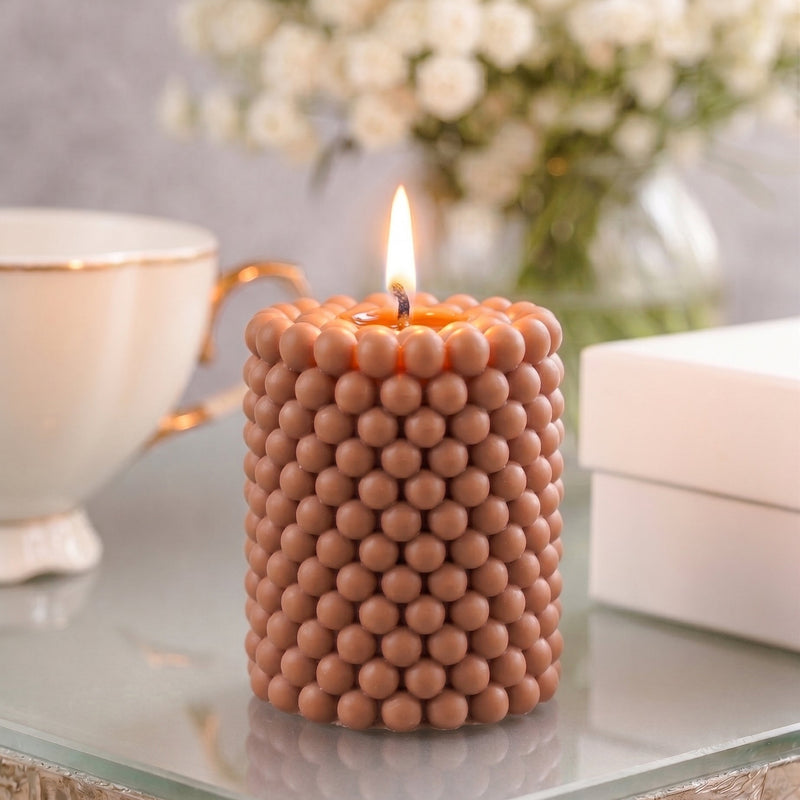 A lit cylindrical candle with a textured, beaded surface in a warm terracotta-brown color, styled on a glass table with a white teacup, white boxes, and a blurred bouquet of white flowers in the background.