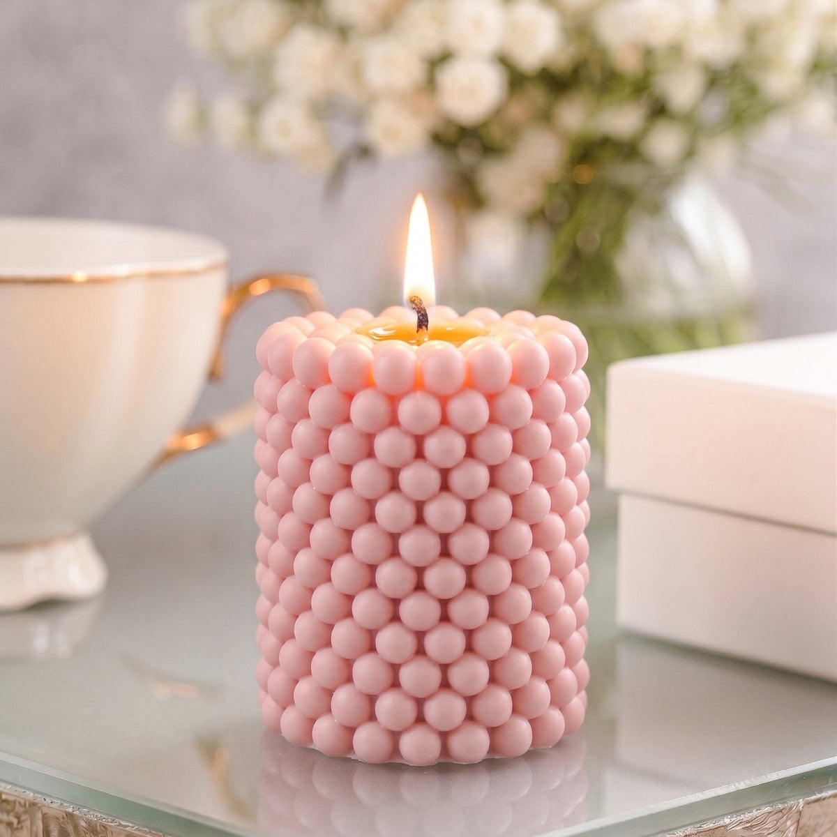 A lit pink candle with a textured, beaded surface sits on a glass table in a styled setting with a teacup, flowers, and a box in the background.