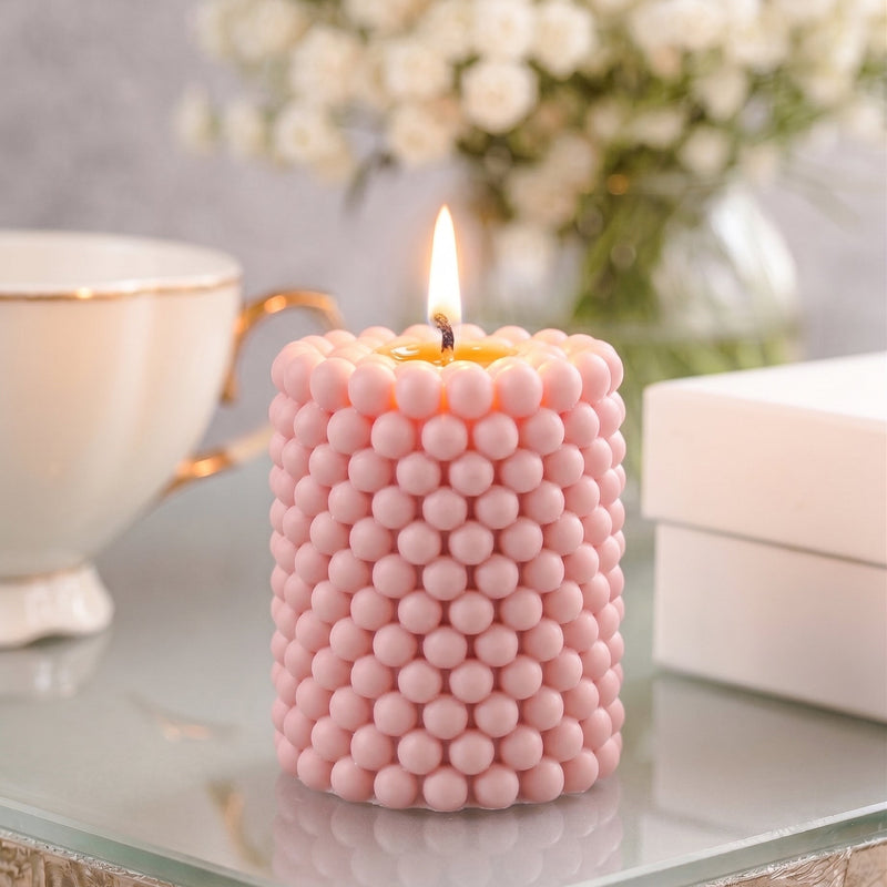 A lit pink candle with a textured, beaded surface sits on a glass table in a styled setting with a teacup, flowers, and a box in the background.