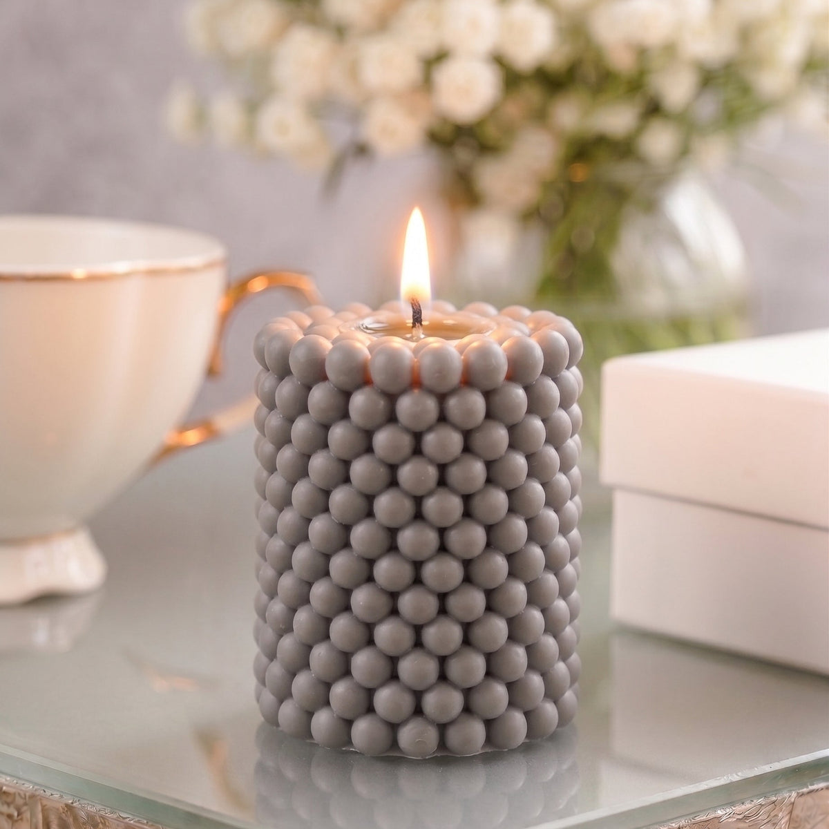 A modern, burning bubble cylinder candle in a sophisticated silver-grey tone, featuring a repetitive, tightly grouped spherical bead design. The candle sits on a polished glass table surface reflecting its form, with a soft-focus background displaying a luxury porcelain teacup, stacked boxes, and a fresh white floral arrangement.