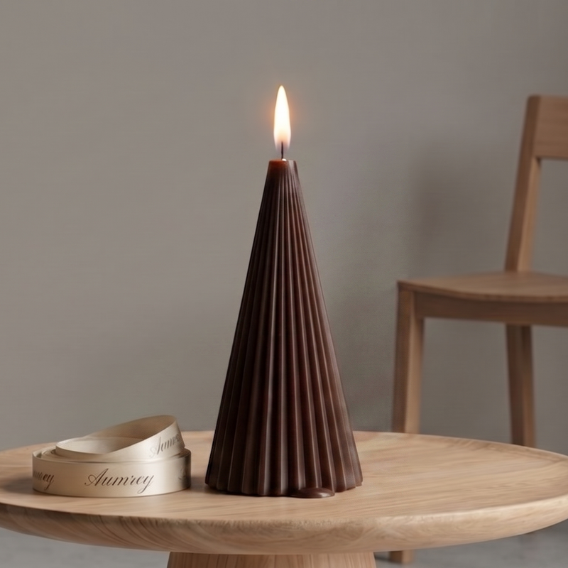 A burning, cone-shaped structural candle in a dark desert drift brown shade, featuring sharp, vertical accordion-style fluting. The candle is placed on a round wooden pedestal table alongside a coiled, elegant cream satin ribbon printed with the luxury brand name "Aumrey" in cursive script. A minimalist wooden chair is softly blurred against a plain neutral background wall.