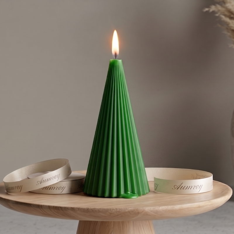 A decorative cone candle with a steady burning flame, colored in a vibrant earthy green or holiday pine green. The conical wax body is molded with clean vertical pleated ridges and displayed on a light wood tabletop stand. An uncoiled ivory silk ribbon with the branding "Aumrey" surrounds the base, with a rustic textured ceramic vase holding fluffy pampas grass stalks in the background.