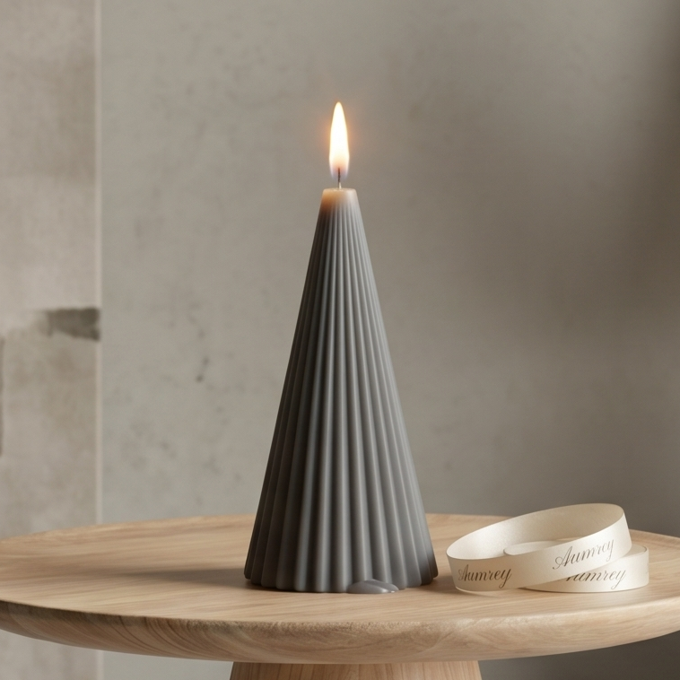A burning, minimalist cone pillar candle in a sophisticated silver-grey or slate-grey finish, defined by precise, vertical fluted pleats. The candle rests on a smooth wooden display tray next to an elegantly rolled ivory brand ribbon reading "Aumrey". The neutral lifestyle backdrop features an artisan stone vase filled with dried ornamental lagurus and pampas grass.
