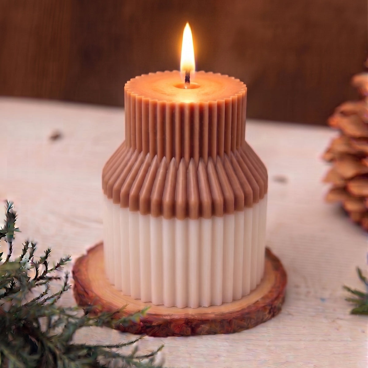 A two-tiered candle with a warm brown top section and an off-white bottom section, styled on a rustic wooden slice coaster with pine branches and a pinecone in the background.
