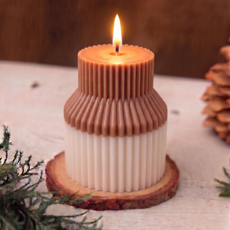 A two-tiered candle with a warm brown top section and an off-white bottom section, styled on a rustic wooden slice coaster with pine branches and a pinecone in the background.