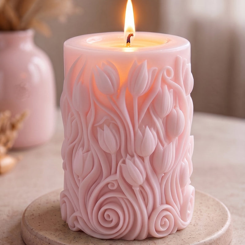 A pink cylindrical candle with a lit flame, featuring a raised floral tulip pattern on the front, displayed on a round beige coaster on a tabletop.