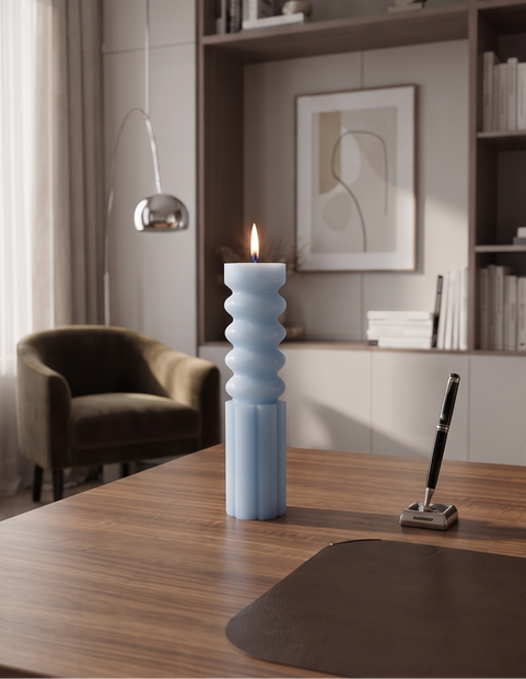 A modern interior scene showing a wooden desk in a home office with a light blue ribbed candle holder candle, a dark brown desk mat, shelving, chairs, and a window with sheer curtains.