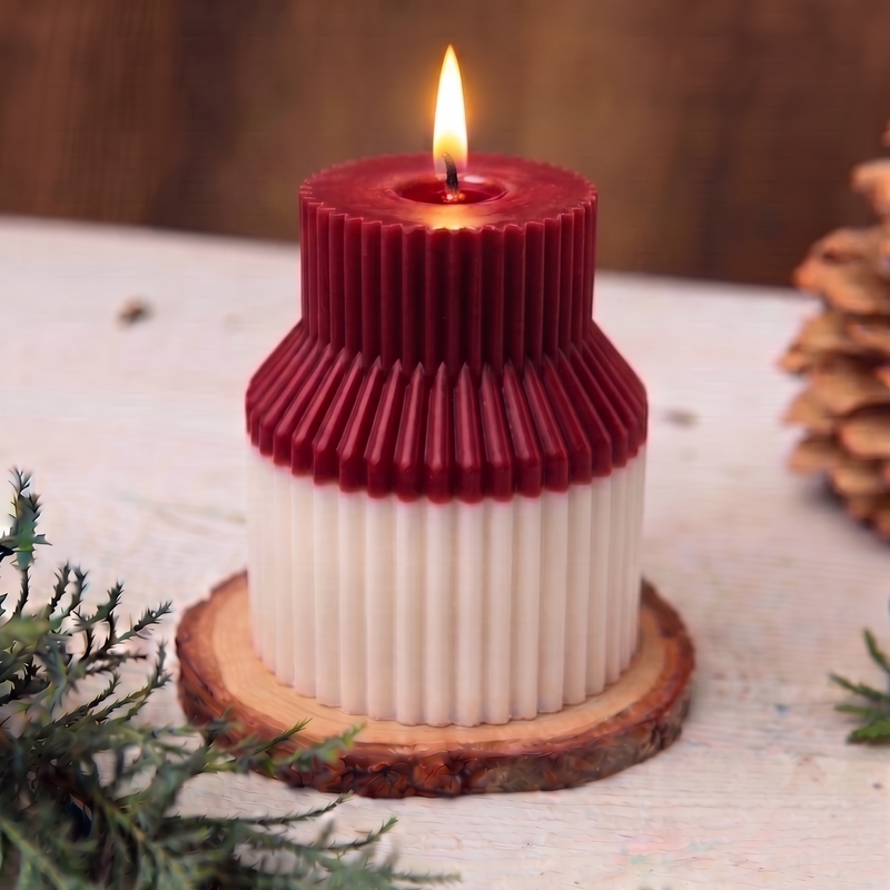 A two-toned ribbed pillar candle with a dark red top and a white bottom, lit with a small flame and displayed on a wood slice coaster with greenery in the foreground.