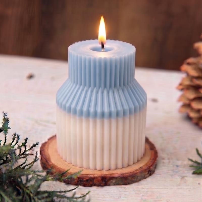 A two-toned candle with a light blue top and a cream-colored bottom, styled on a rustic wooden slice coaster with green pine branches in the foreground and a pinecone in the background.