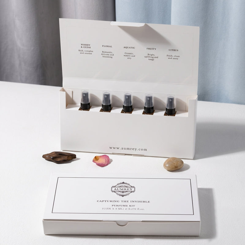 A boxed fragrance oil set displayed on a white surface with a soft gray curtain backdrop. The set includes an open white box holding six small dark glass bottles, a closed matching box in front, and a few small decorative stones and a rose petal nearby.