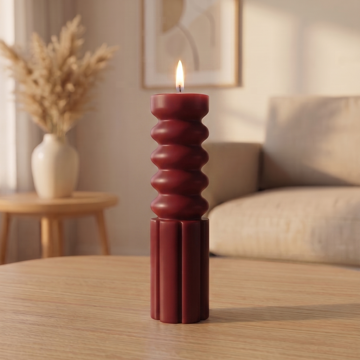 A dark red ribbed pillar candle with a lit flame, standing on a light wood table in a softly lit living room.