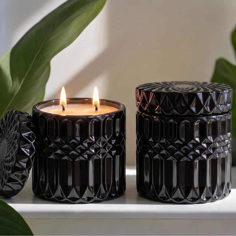 Two black textured glass jar candles on a white shelf, with one candle open and lit showing a warm flame, surrounded by green leaves against a soft white background.