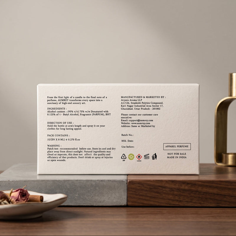 The back of a white perfume box is shown on a dark wooden surface, with product text visible. A brass diffuser and dried flowers sit nearby against a neutral beige background.