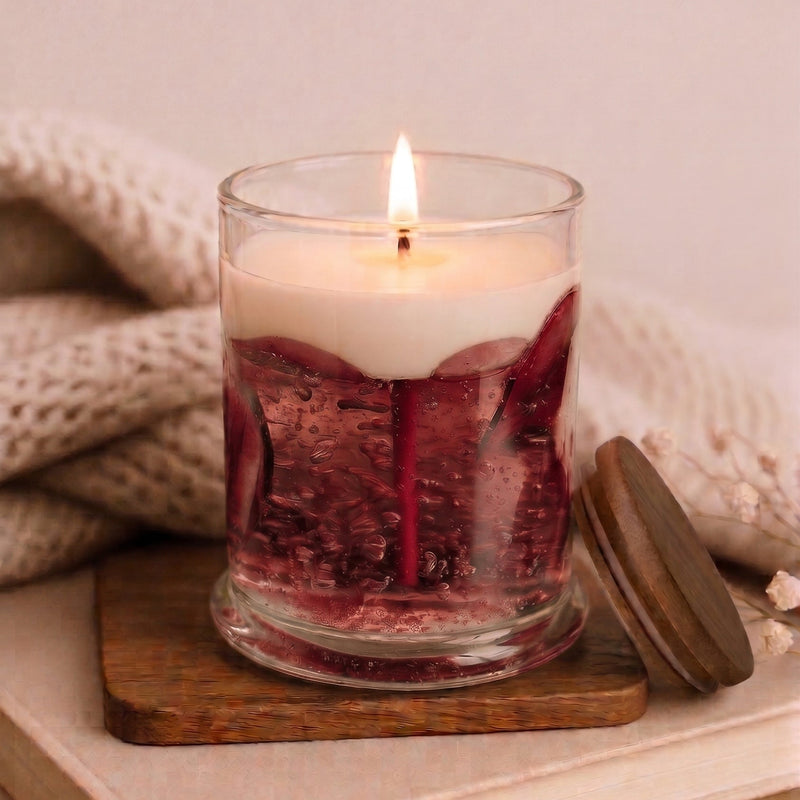 A lit candle in a clear glass jar with a cream-colored wax top and deep red wax bottom, styled on a wooden block over stacked books with a knitted blanket in the background.