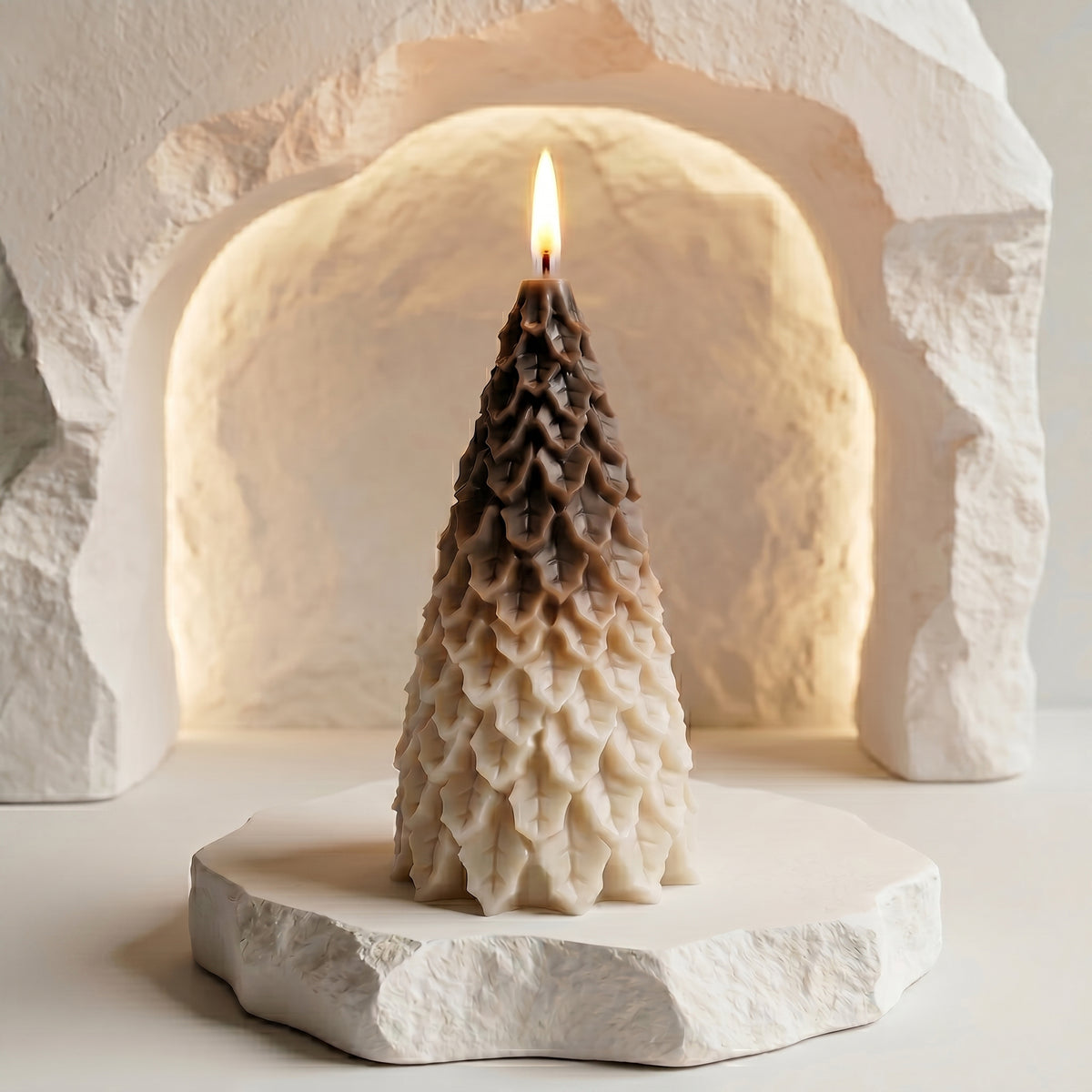 A lit festive tree candle with a detailed, layered pinecone or foliage texture, featuring an ombré color transition from a warm, earthy desert drift brown at the top to a cream-white base. The candle is placed on a rough-edged white stone platform, centered within a contemporary, textured stone archway backlit with a soft, warm glow.